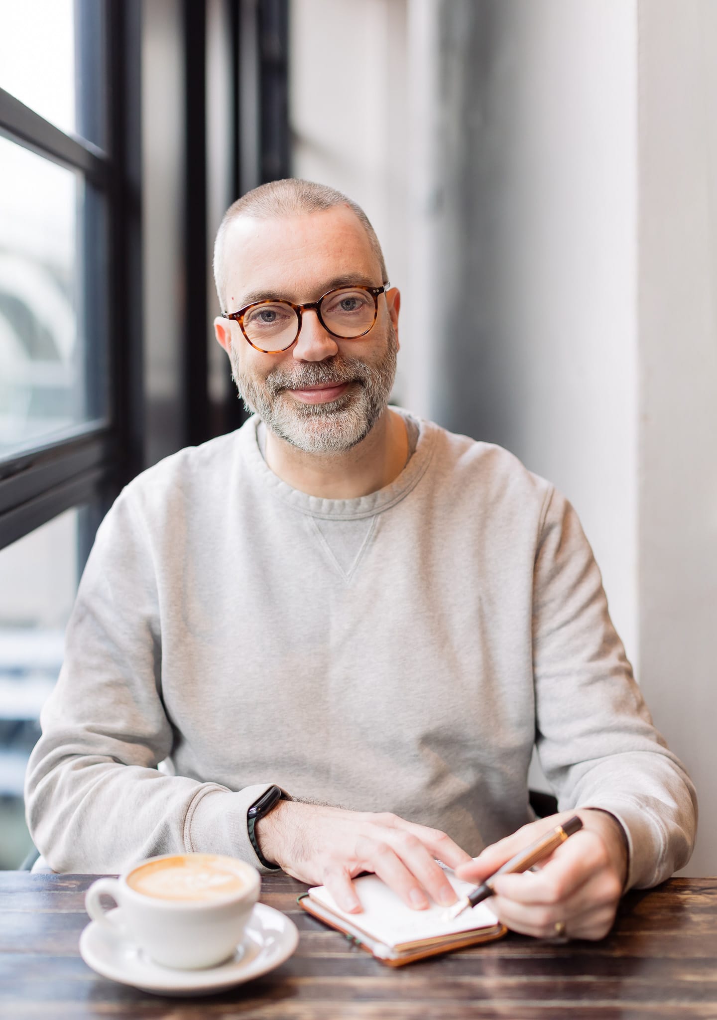 Anthony Gladman, drinks writer and journalist, sits at a wooden café table by a large window, writing in a small notebook with a fountain pen beside a coffee in a white cup and saucer.
