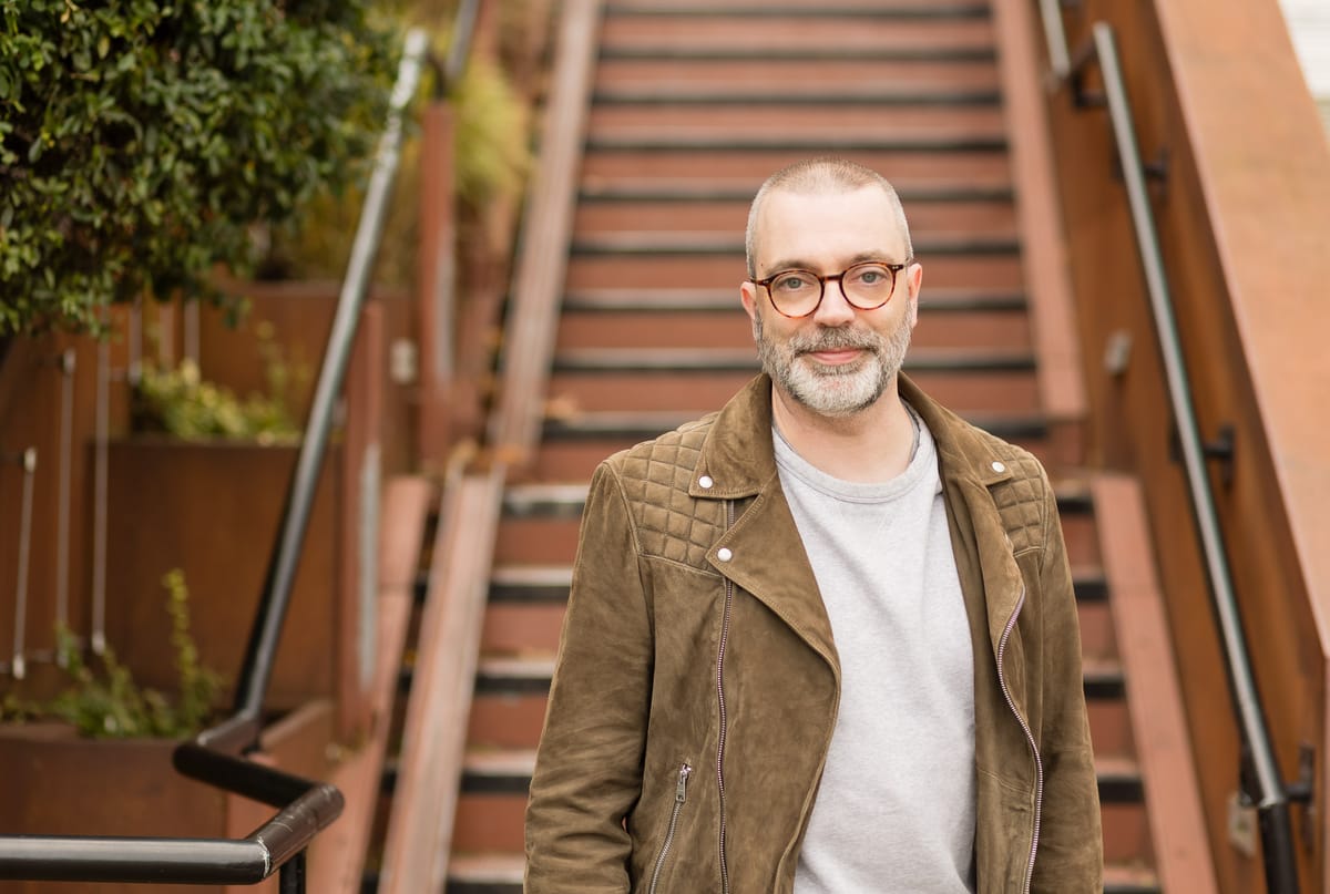 A white man in his 40s with cropped salt-and-pepper hair and beard, glasses, brown suede biker jacket, and grey sweatshirt, standing by a rusted iron staircase with plants.