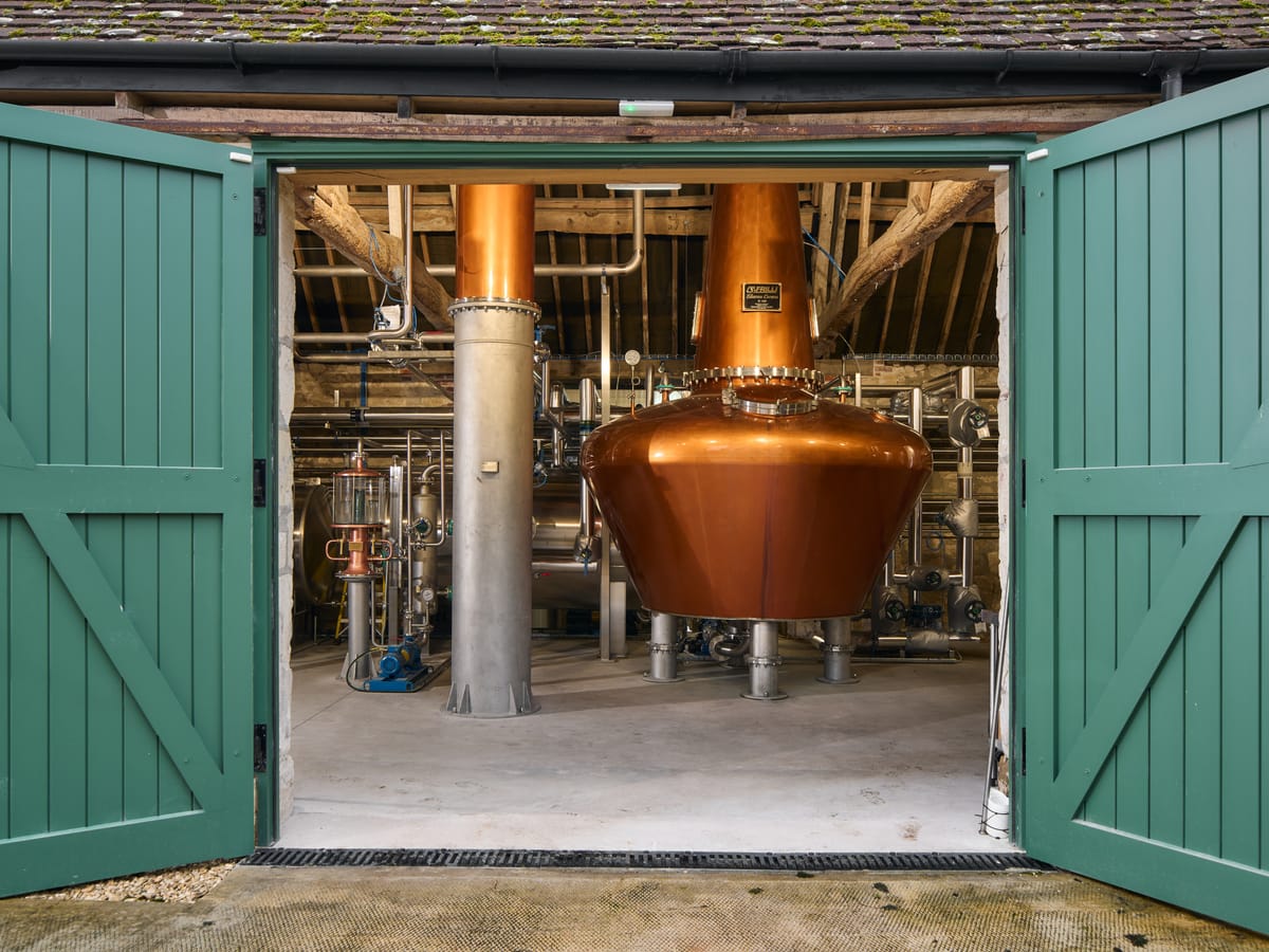 A copper pot still viewed through the open wooden doors of a converted stone barn. The doors are painted sage green. There is neat and organised pipework on the wall behind the still.