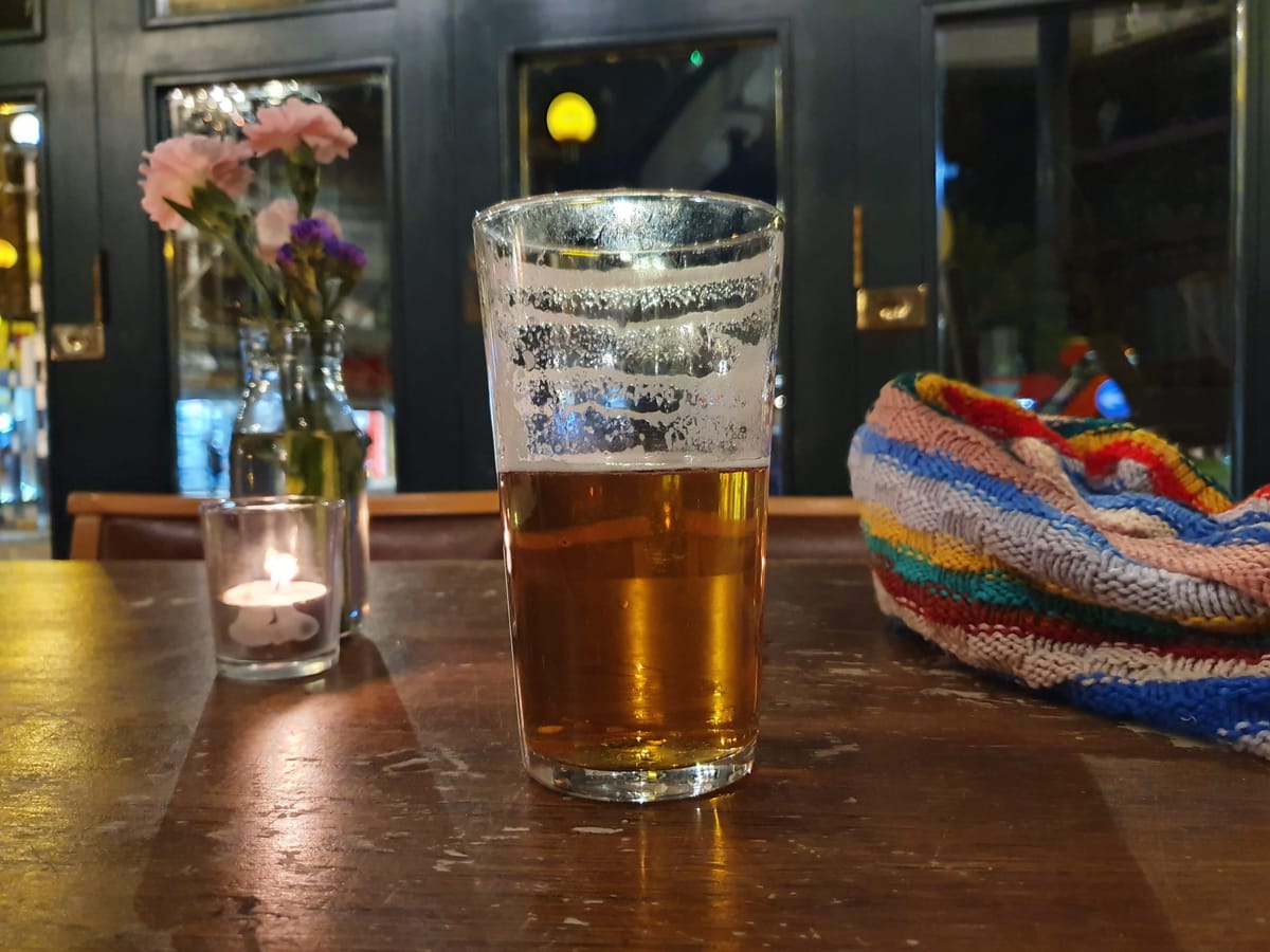 Half-full pint glass of amber beer on a worn wooden table in a cosy pub, with a lit tealight candle, small vase of pink and purple flowers, and a colourful knitted scarf nearby.