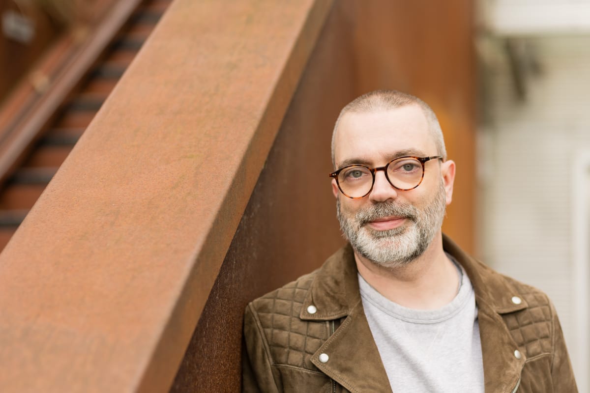 Anthony, in a brown suede biker jacket, leans against a rust-coloured staircase