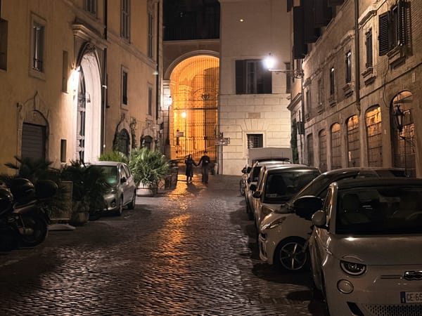 Narrow cobblestone street at night with parked cars and motorbikes, flanked by tall, shuttered buildings. A glowing archway illuminates two distant pedestrians under warm streetlights.